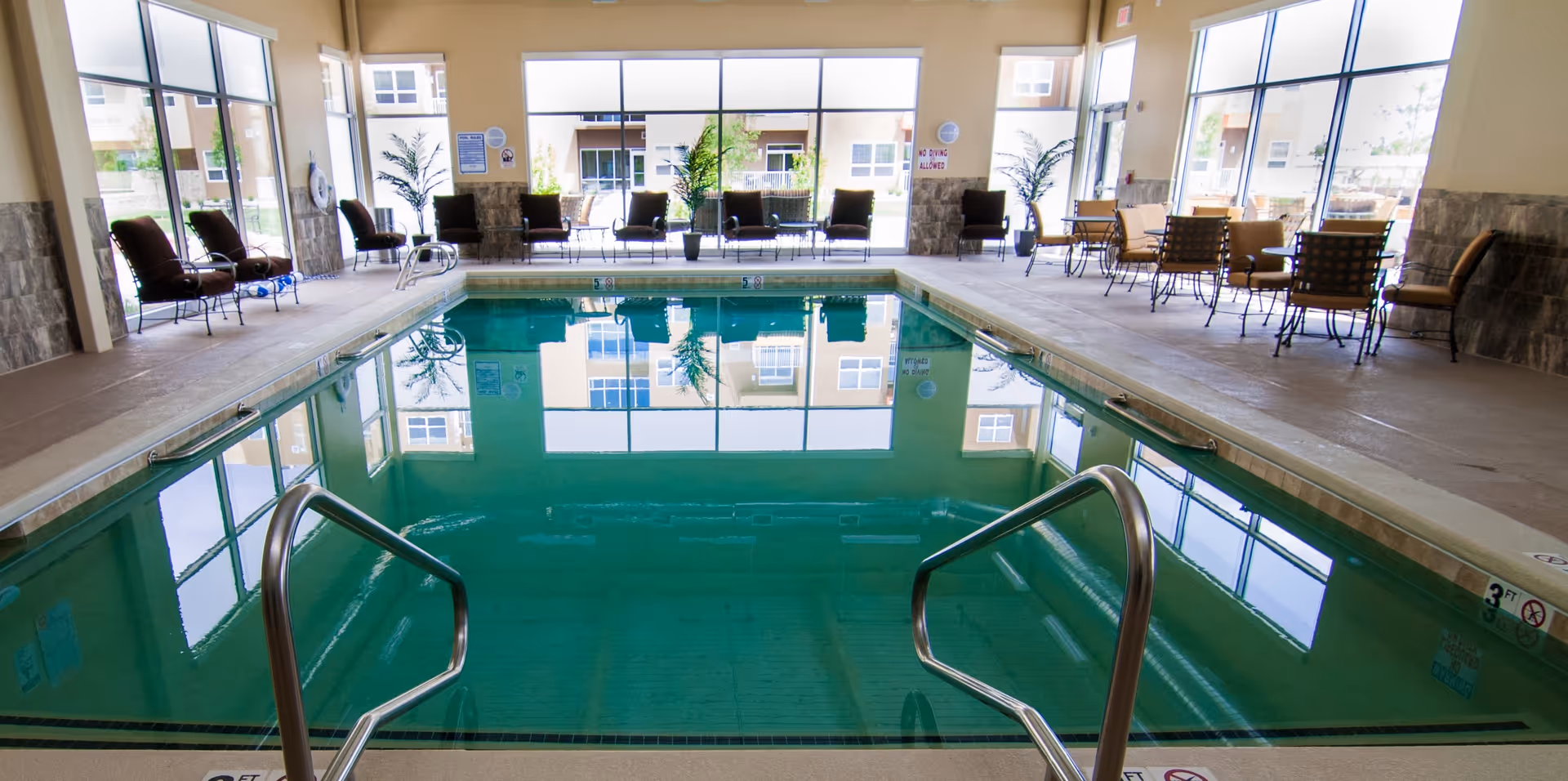Indoor swimming pool with handrails, lounge chairs and tables along large windows reflecting in the water.