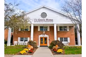 Front exterior view of The Green Home facility, a two-story brick building with white trim and a triangular pediment above the entrance. The entrance has double wooden doors with glass panels, flanked by white columns. There are flower beds with colorful flowers and shrubs on either side of the walkway leading to the entrance. Trees with sparse leaves are visible on both sides of the building.