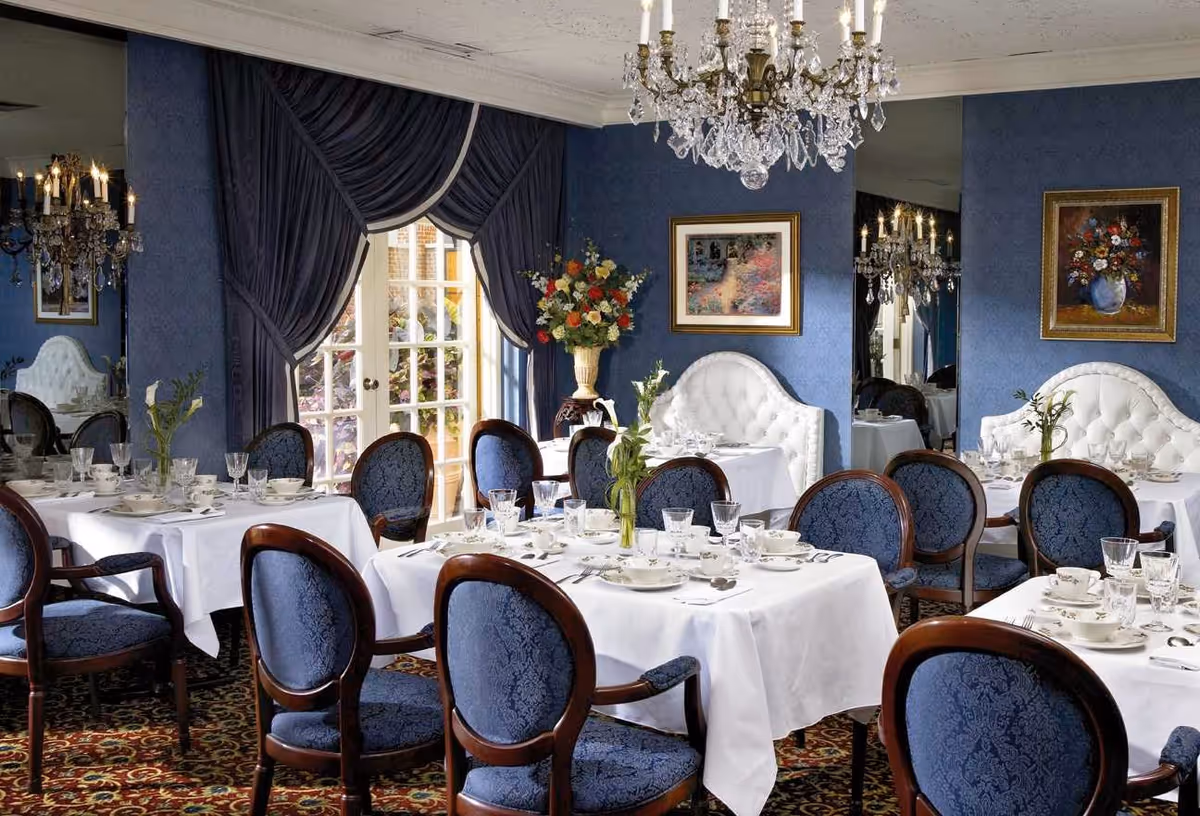 Elegant dining room with tables covered in white tablecloths, set with fine china, glassware, and silverware. The room features blue upholstered chairs with wooden frames, blue walls with framed artwork, a large floral arrangement, and a crystal chandelier hanging from the ceiling. French doors with dark blue drapes lead outside.