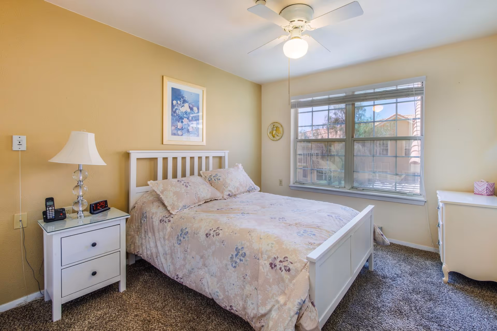 A cozy bedroom with a white wooden bed frame and floral bedding. There is a white nightstand with a lamp, a cordless phone, and a digital alarm clock showing 11:29. A large window with blinds lets in natural light, and a ceiling fan with a light is on the ceiling. The walls are painted a warm beige color, and there is a framed floral picture hanging above the bed.