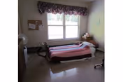 Small nursing-home bedroom with a single bed covered by a pink-striped blanket under a window, a bedside table with a lamp, a corkboard on the wall, and a fan and walker in the room.