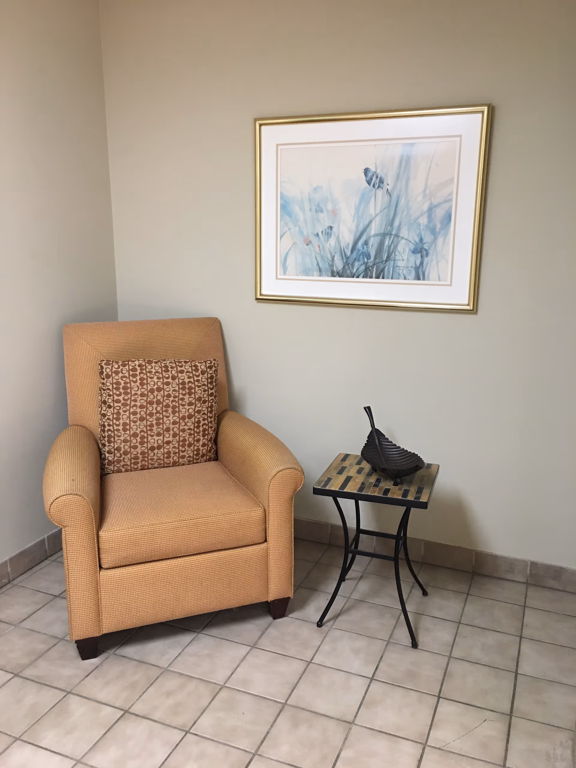 A cozy corner with a cushioned armchair featuring a patterned pillow, a small side table with a decorative black bowl, and a framed artwork of blue flowers hanging on a beige wall. The floor is tiled with light-colored square tiles.