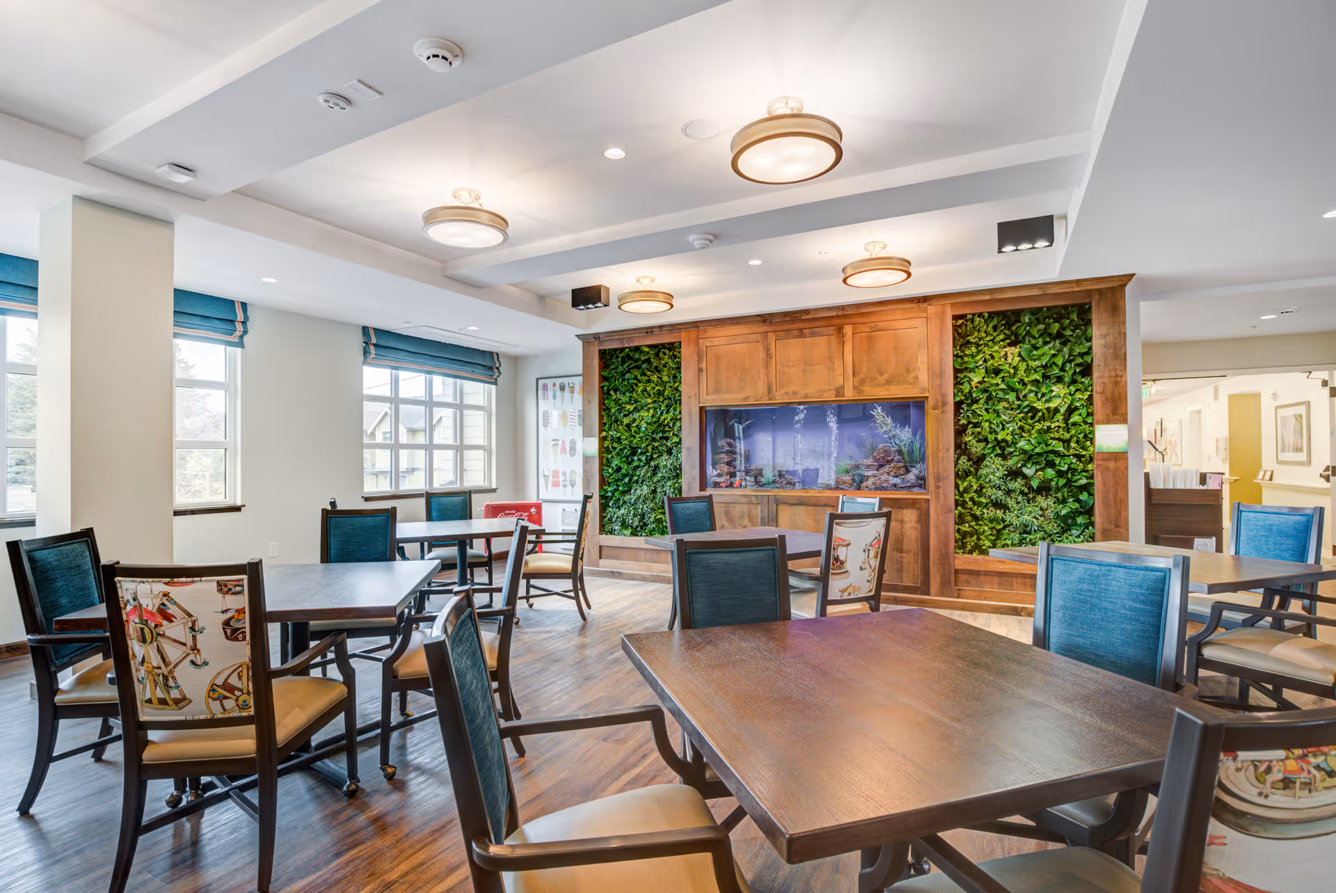 A bright and spacious dining room with several wooden tables and chairs. The room features large windows with blue Roman shades, a wooden wall unit with a built-in aquarium, and vertical green plant panels on either side of the aquarium. The ceiling has modern round light fixtures, and the floor is a polished wood finish.