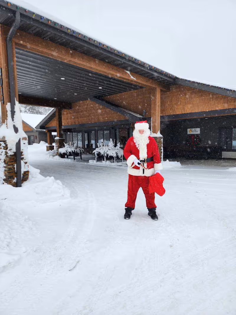 A person dressed as Santa Claus stands in the snowy driveway in front of a covered entrance to a building.