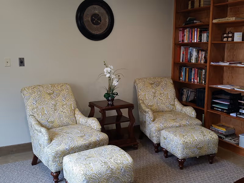 A cozy sitting area with two patterned armchairs and matching ottomans, separated by a small wooden side table with a white orchid plant. A large wooden bookshelf filled with books and papers is on the right wall, and a round wall clock hangs above the chairs on a beige wall.