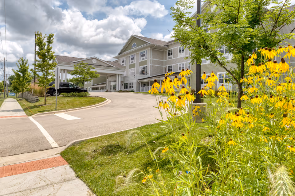 Exterior view of Cedarhurst Senior Living of Naperville showing a three-story building with a covered entrance driveway. The foreground features a garden with yellow flowers and green trees under a partly cloudy sky.