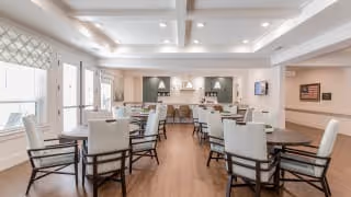 A spacious and well-lit dining room with multiple tables and cushioned chairs arranged neatly. The room features a coffered ceiling with recessed lighting, large windows with patterned valances allowing natural light, and a serving area with a countertop and cabinetry at the far end. The flooring is wood, and the walls are painted in neutral tones with some decorative elements.