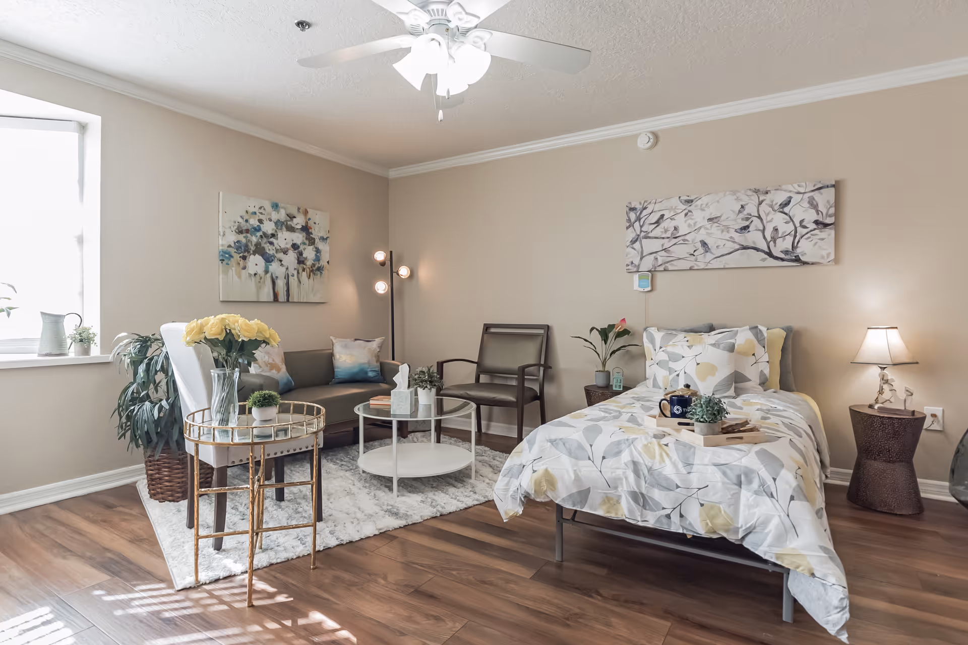 A cozy and well-lit bedroom in a senior living facility named The Pointe, featuring a single bed with floral bedding, a small side table with a lamp, a sitting area with a sofa, armchair, and round glass coffee table on a white rug, decorative plants, and wall art depicting flowers and birds. The room has wooden flooring and a ceiling fan with lights.