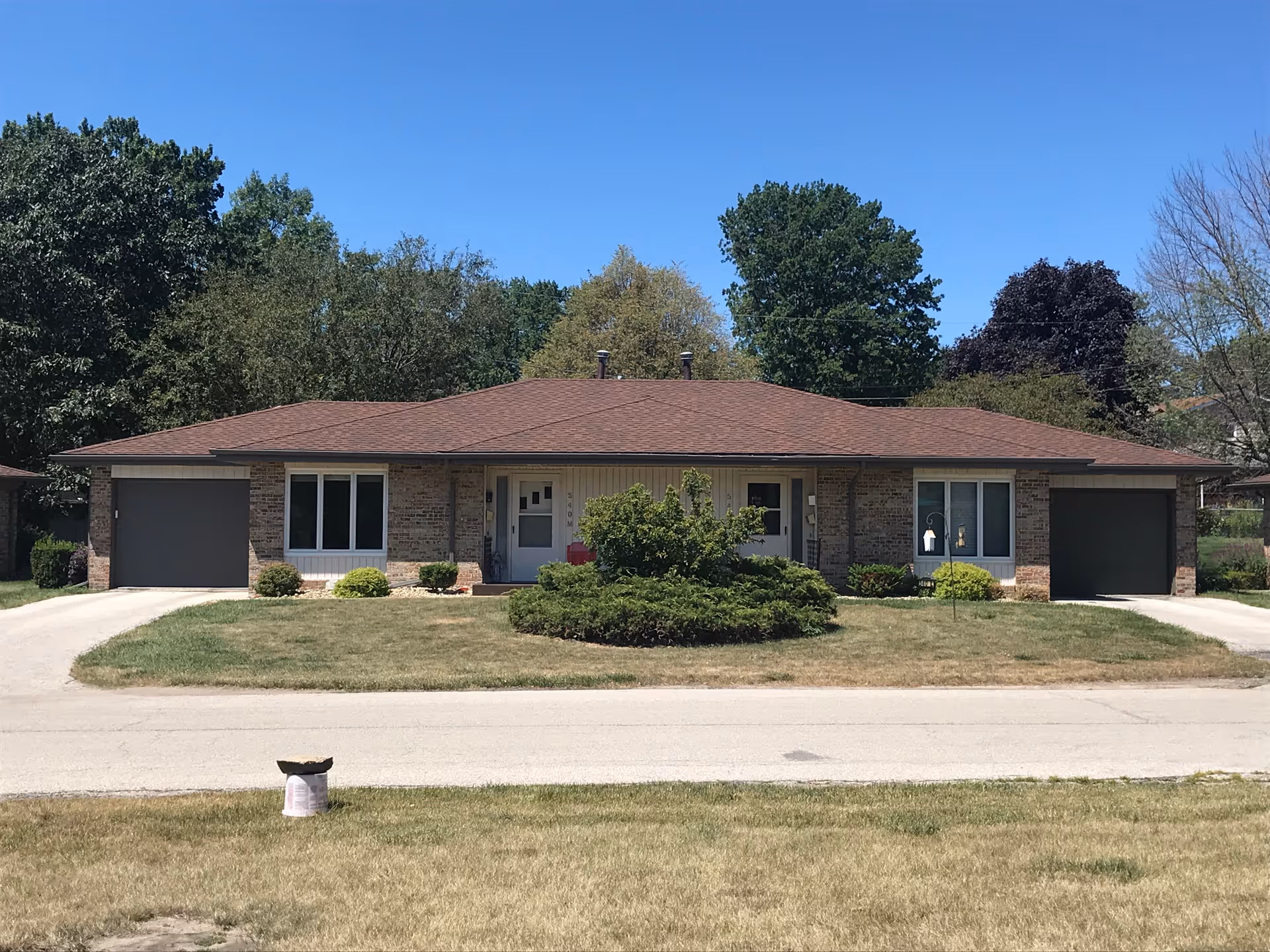 A single-story brick building with a brown shingled roof, two garages on either side, and a central entrance. There is a small circular shrubbery in front of the building, surrounded by a lawn and a paved driveway. Trees are visible in the background under a clear blue sky.