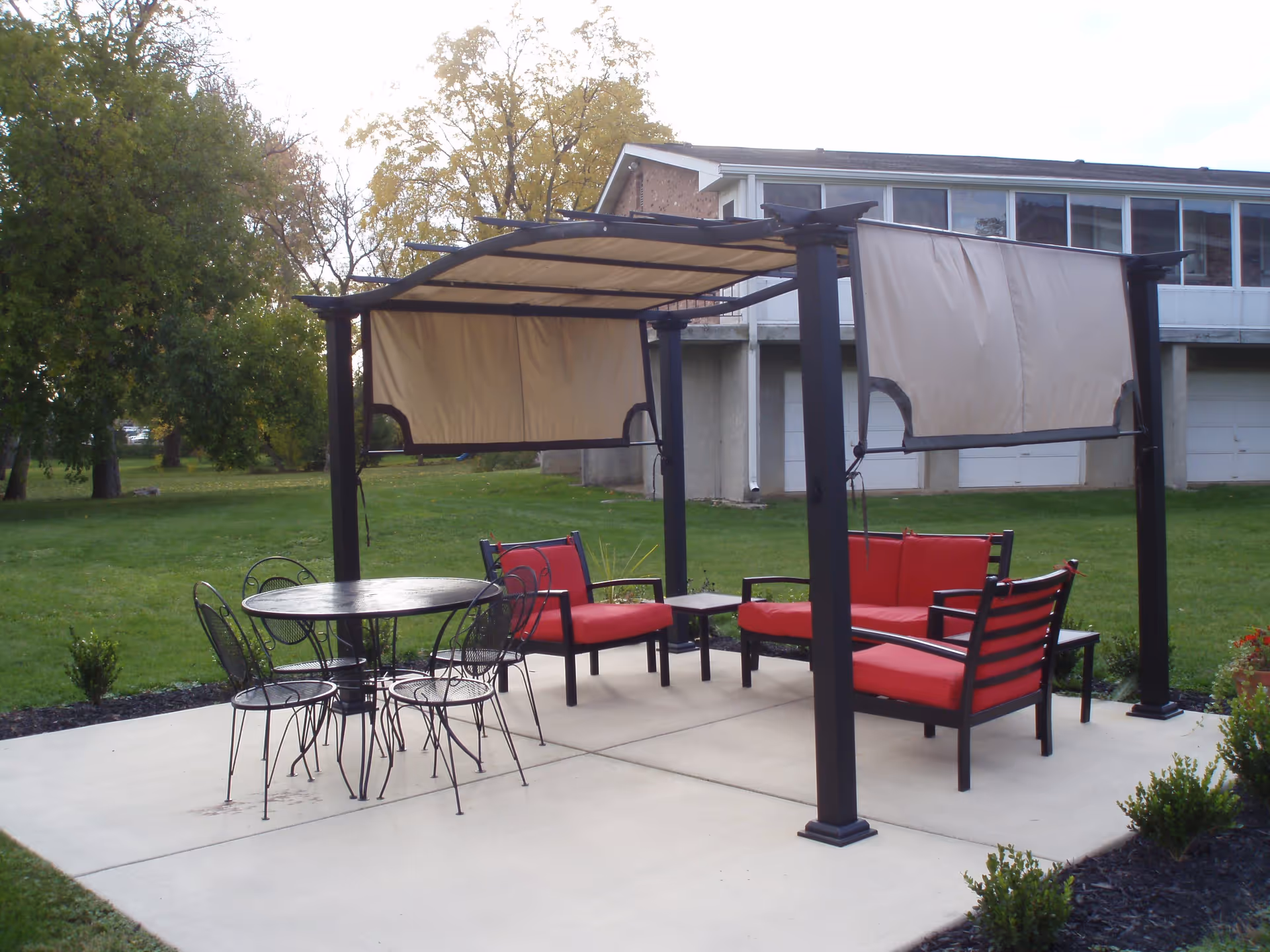 Outdoor patio area with a metal pergola featuring beige fabric shades. Underneath are two red cushioned benches, a small table, and a round black metal table with four matching chairs. The patio is surrounded by green grass and trees, with a building visible in the background.