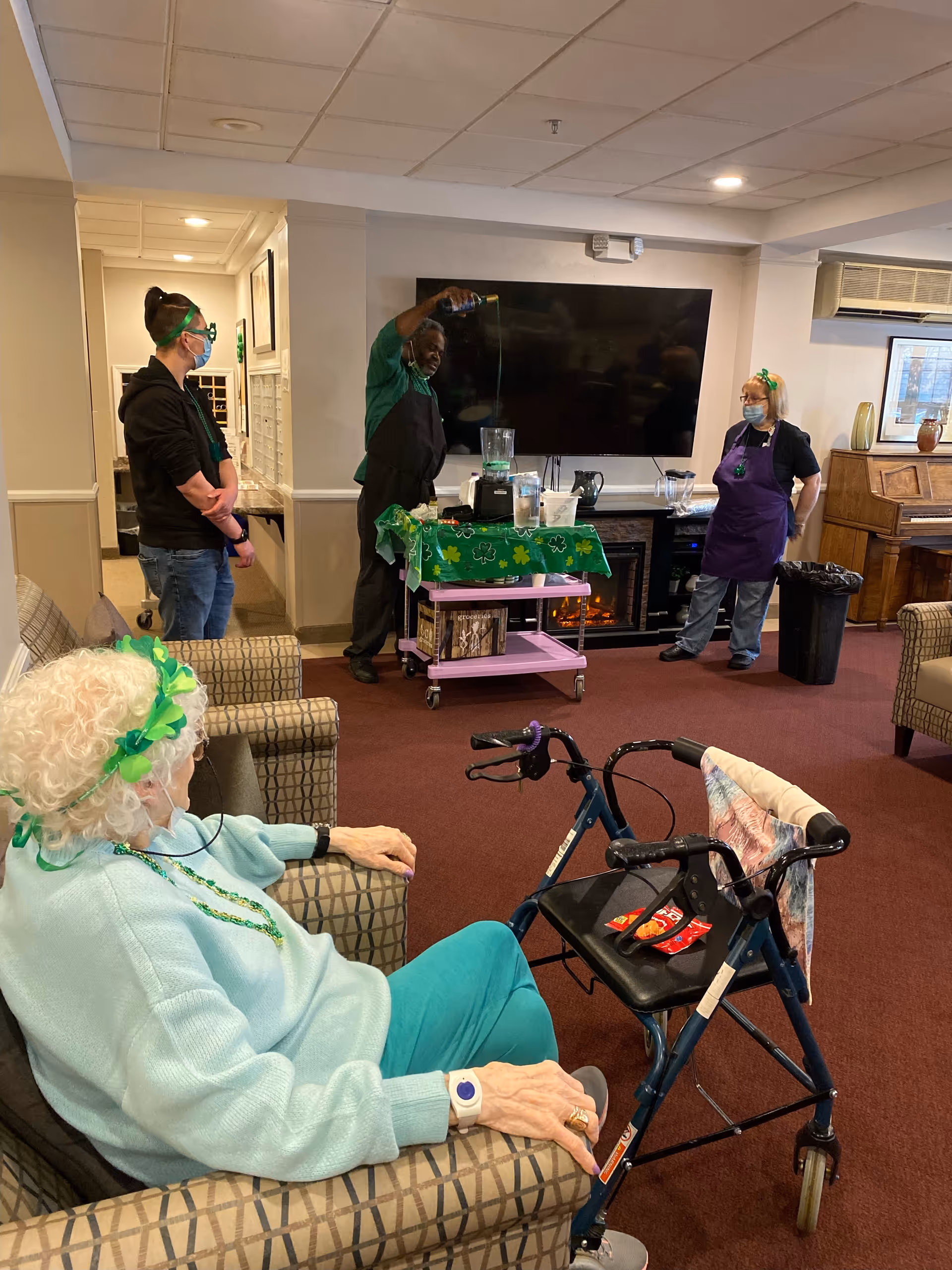 Residents and staff gathered in a senior living common room while a staff member pours a drink from a blender at a holiday-themed activity.