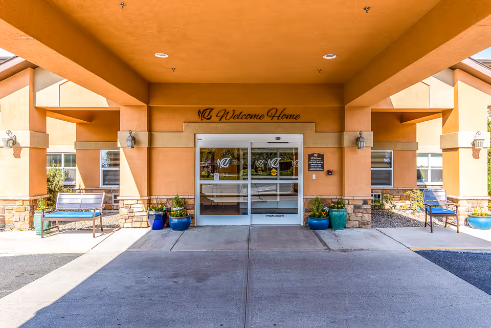 Covered front entrance with a 'Welcome Home' sign above sliding glass doors, benches and potted plants under a portico.