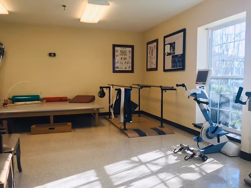 A therapy or rehabilitation room with parallel bars for walking exercises, a stationary exercise bike near a large window, a padded therapy table with various therapy tools on it, and anatomical charts on the walls.