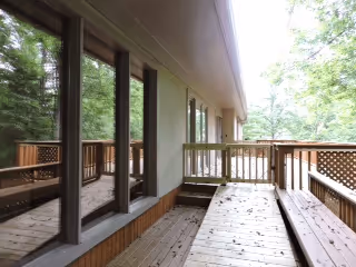 Wooden deck attached to a building with large glass windows reflecting trees, surrounded by a wooden railing and lush green trees in the background.