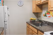 Small kitchen area with a sink, granite countertop, wooden cabinets, refrigerator, wall clock, and a bowl of fruit.