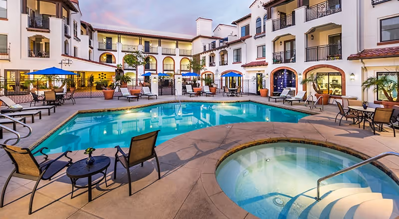 Outdoor swimming pool and hot tub area surrounded by a multi-story building with balconies and arched windows, patio tables with blue umbrellas, and lounge chairs.