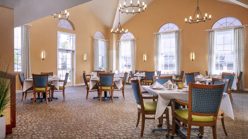 A spacious dining room with multiple tables covered in white tablecloths, each set with napkins, cups, and small decorative lanterns. The room features tall arched windows with patterned curtains, beige walls, and carpeted flooring. Modern chandeliers hang from the high ceiling, providing warm lighting.
