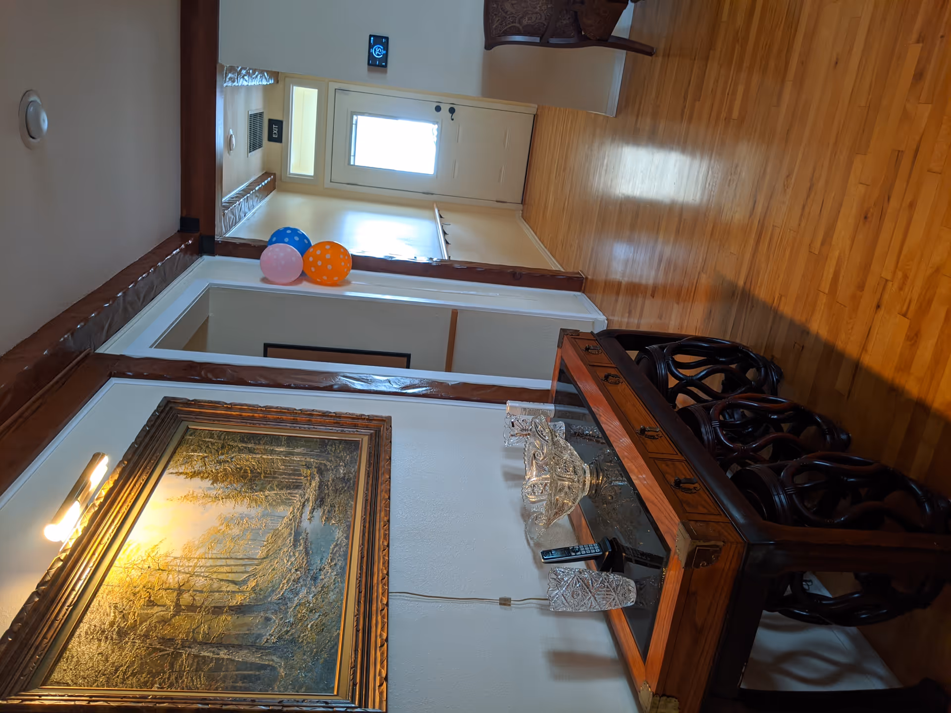 A hallway with wooden flooring and white walls, featuring a wooden console table with glass decorative items and a cordless phone. Above the table is a framed painting of a forest scene illuminated by a wall light. There are colorful balloons attached to the wall near a doorway, and a door with a window at the end of the hallway. A thermostat is mounted on the wall to the right.