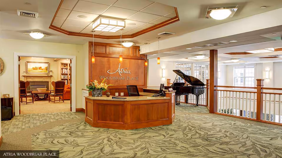 Reception area of Atria Woodbriar Place featuring a wooden front desk with a flower arrangement, a sign on the wall behind the desk, a grand piano in the background, and a seating area with chairs and a fireplace visible through an open doorway.