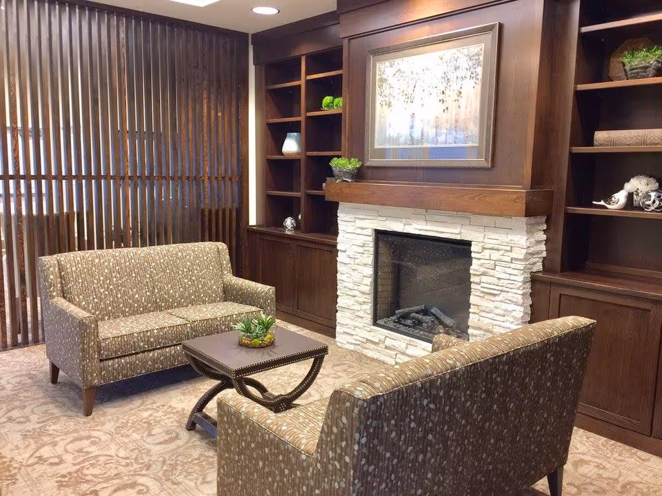 A cozy sitting area with two patterned sofas facing each other across a small wooden coffee table with a plant centerpiece. Behind one sofa is a modern fireplace with a white stone surround and a wooden mantel, above which hangs a framed artwork. Flanking the fireplace are built-in dark wood shelves with decorative items. Vertical wooden blinds cover the window on the left side of the room.