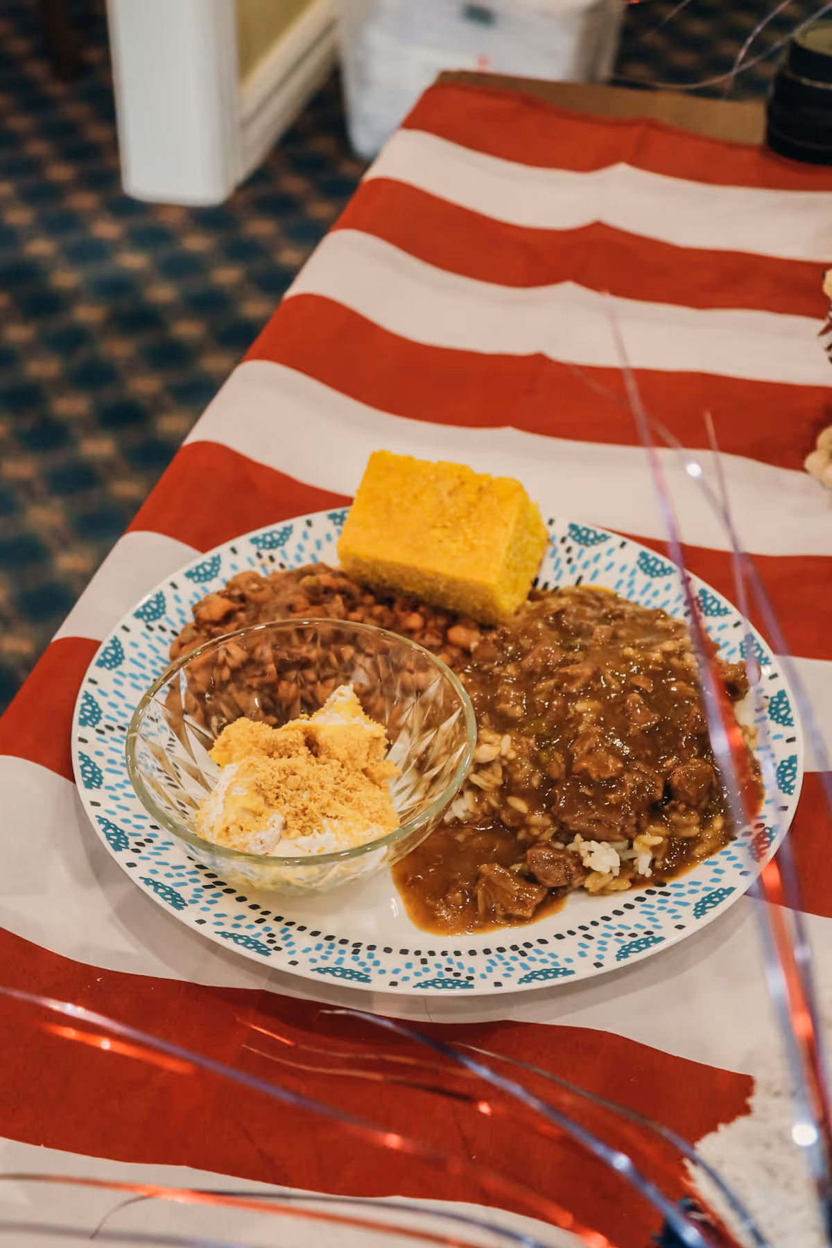 A plate of food on a red and white striped tablecloth, featuring a serving of cornbread, black-eyed peas, a meat and gravy dish over rice, and a small glass bowl with a dessert topped with crumbled topping.