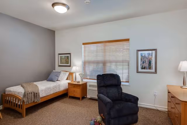 A tidy senior living bedroom with a bed, recliner, dresser, and window blinds.