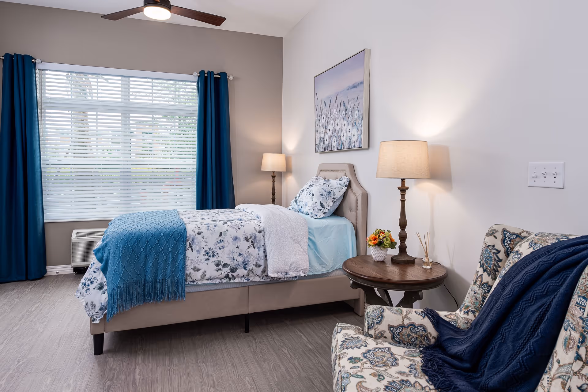 A cozy bedroom with a single bed dressed in floral bedding and a blue throw blanket. The room features a large window with blue curtains, two bedside lamps on either side of the bed, a round wooden side table with a small flower arrangement, and a patterned armchair with a dark blue throw draped over it. The walls are painted white and light gray, and there is a floral painting hanging above the bed.