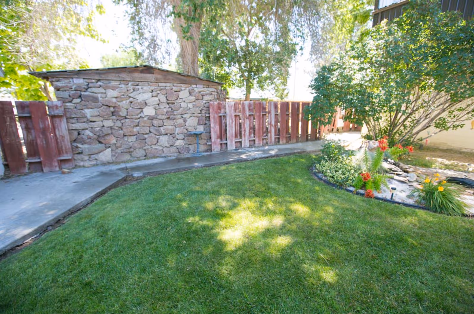 A well-maintained outdoor garden area with green grass, a stone wall, a wooden fence, a concrete pathway, and various plants and flowers including a bush with red flowers and a small pond.