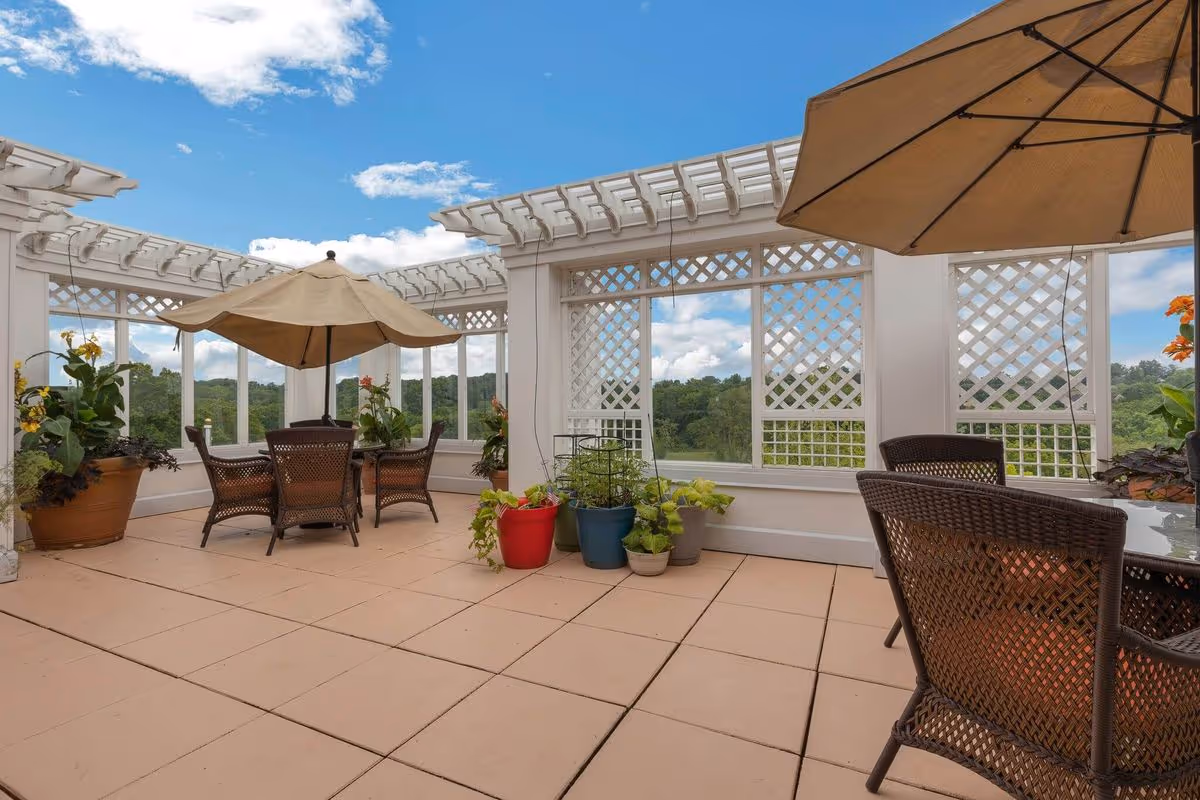 Outdoor patio area with beige tiled flooring, white lattice walls, and a pergola overhead. There are two sets of brown wicker chairs around glass-top tables, each shaded by large beige umbrellas. Several potted plants are placed around the patio, and a scenic view of green trees and a partly cloudy blue sky is visible in the background.