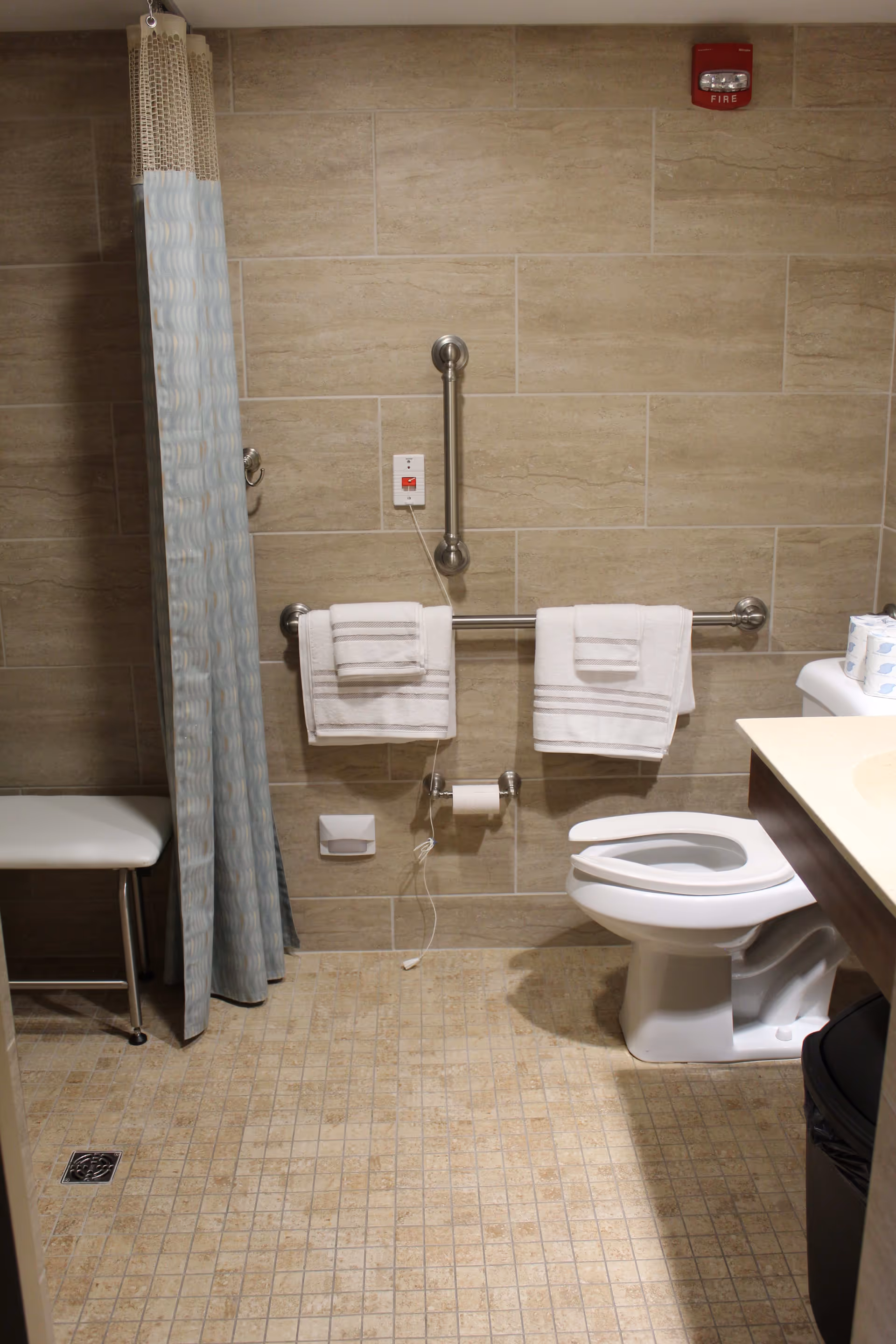 Accessible bathroom with beige tiled walls and floor, a white toilet with an open lid, two towel bars with white towels, a shower curtain partially covering a shower area with a white bench, a fire alarm on the wall, and a trash can next to the sink counter.