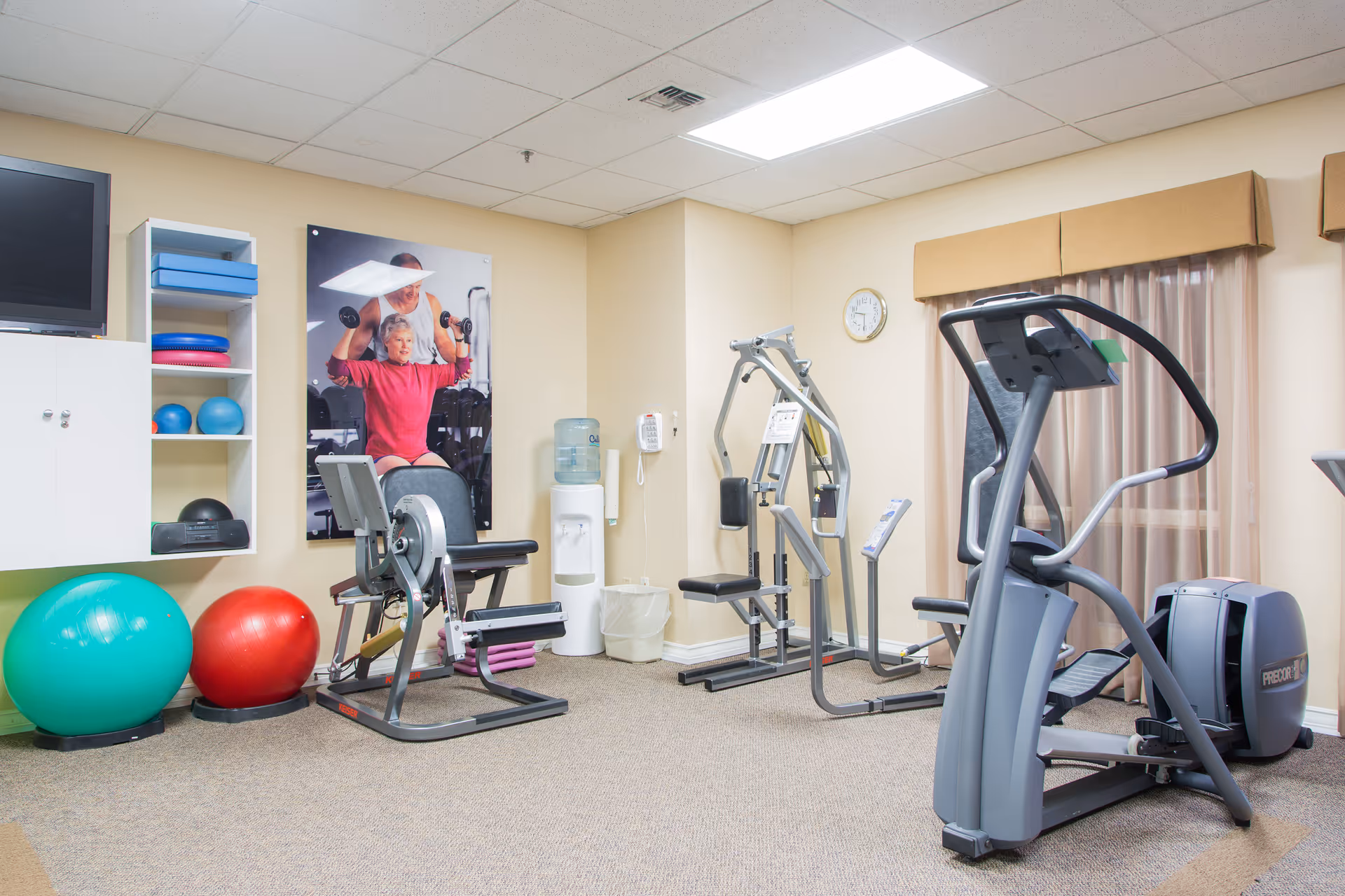 Small fitness room with exercise machines, stability balls, storage shelves and a water cooler.