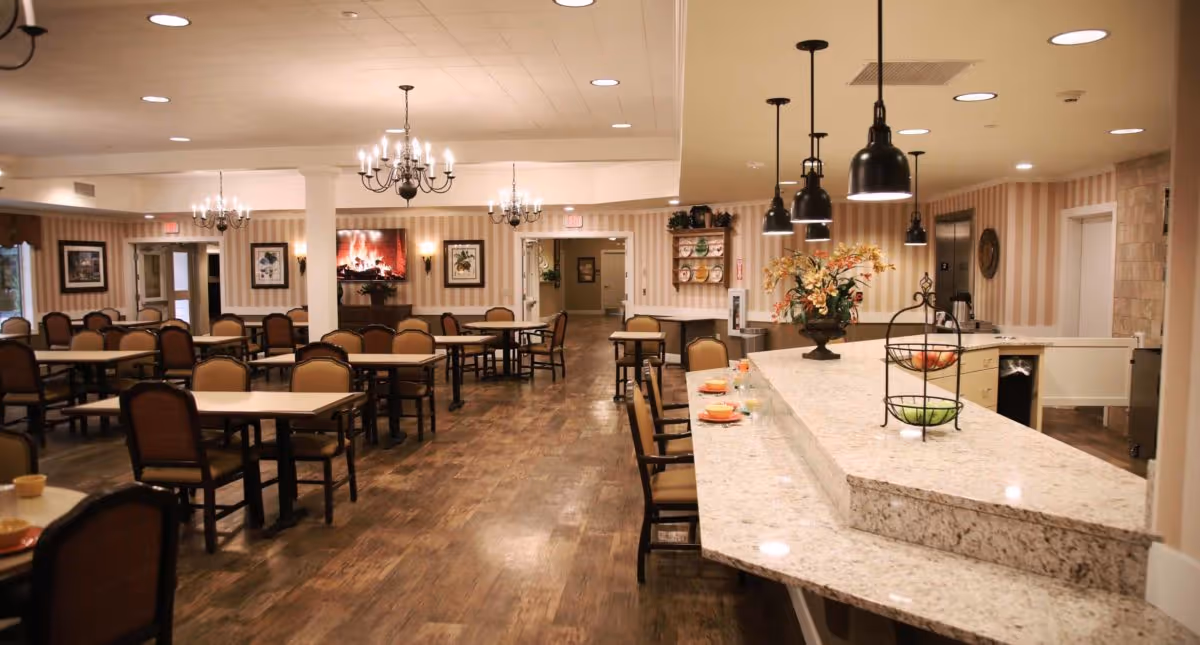 A spacious dining room in a senior living facility with multiple tables and chairs arranged neatly. The room features wooden flooring, chandeliers, and pendant lights hanging over a long granite countertop with chairs. The walls have striped wallpaper and framed artwork, and there is a cozy fireplace in the background.