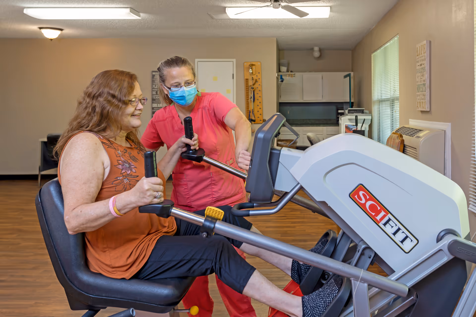 A woman in an orange sleeveless top and black pants is seated on a SciFit exercise machine, using the handles and pedals. A healthcare worker in a pink uniform and blue face mask is assisting her. The room has wooden flooring, beige walls, a ceiling fan, and windows with blinds.