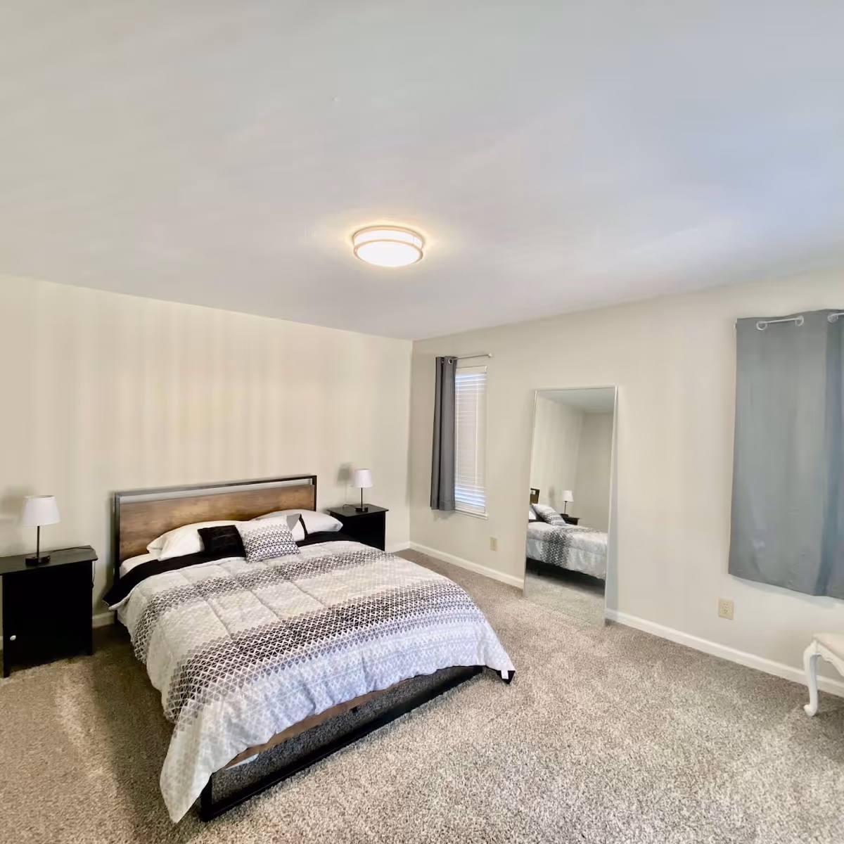 A simple and clean bedroom with a bed featuring a patterned comforter and pillows, two black nightstands with lamps on either side, a large standing mirror leaning against the wall, two windows with gray curtains, and beige carpet flooring.