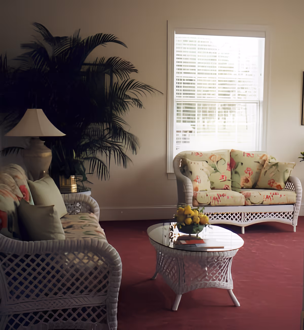A cozy living room area with two white wicker sofas featuring floral cushions, a glass-top wicker coffee table with a bowl of yellow fruit, a large leafy plant, and a window with white blinds letting in natural light.