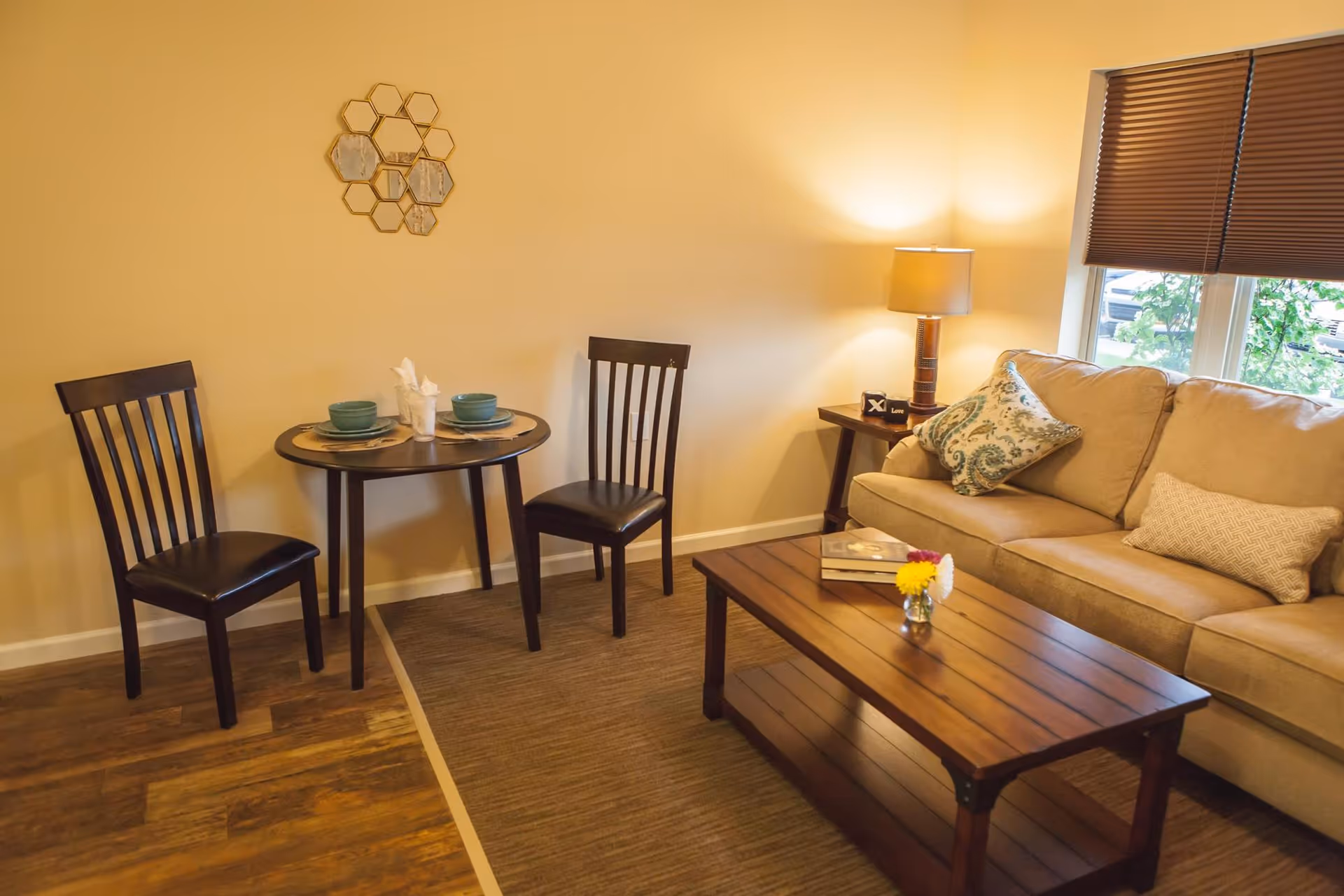 A cozy living room area with a beige couch adorned with patterned pillows, a wooden coffee table with books and a small vase of flowers, a side table with a lamp, and a small round dining table set with two chairs, plates, bowls, and napkins. A window with brown blinds lets in natural light.