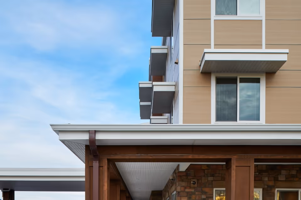 Corner of a modern senior living building showing upper windows, small balconies, and a covered entry with wooden columns against a blue sky.