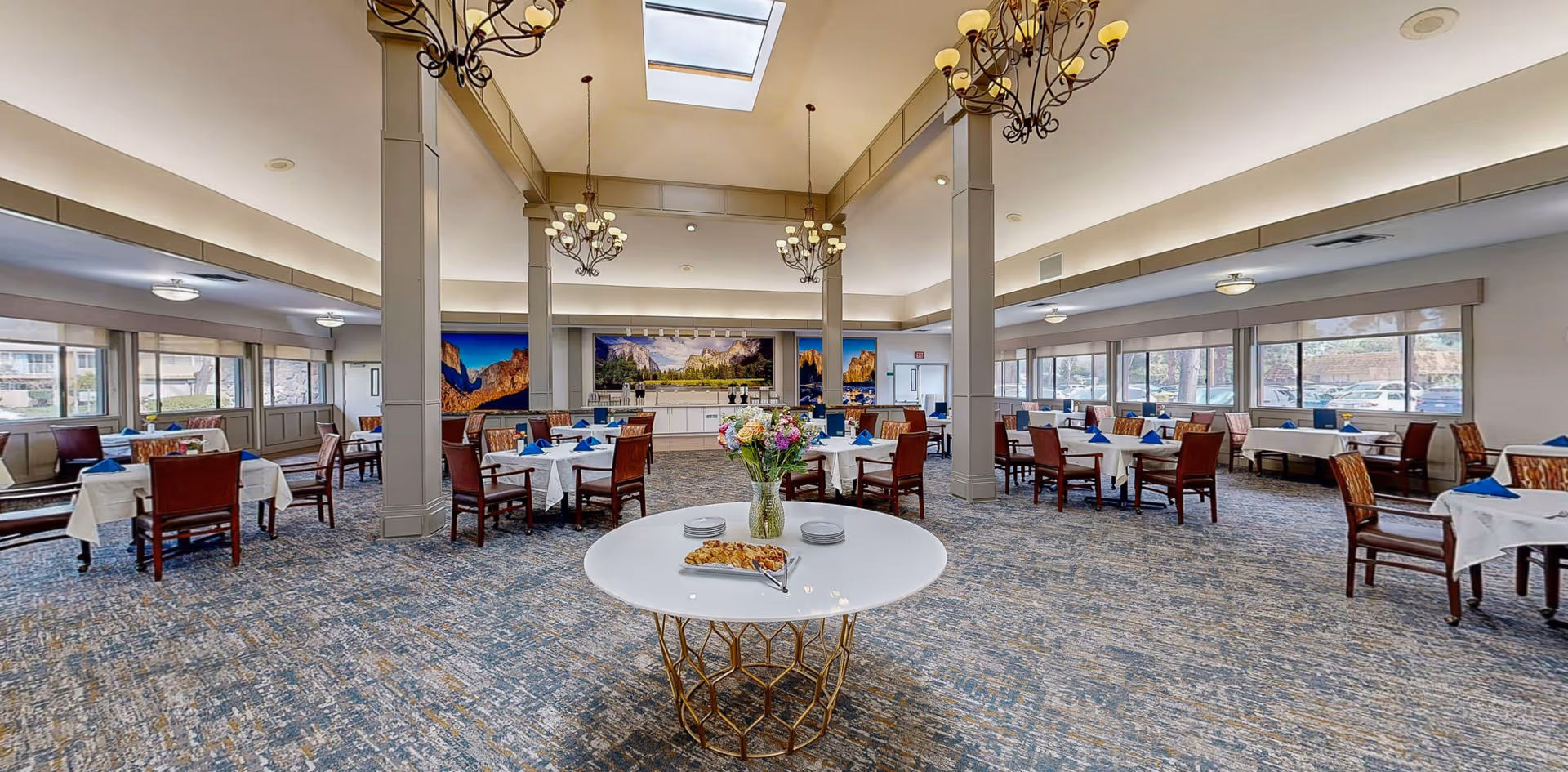 Spacious dining room with multiple tables covered in white tablecloths and set with blue napkins. A round table in the foreground holds a vase of colorful flowers and a plate of pastries. The room features large windows letting in natural light, chandeliers hanging from the ceiling, and scenic landscape artwork on the far wall.