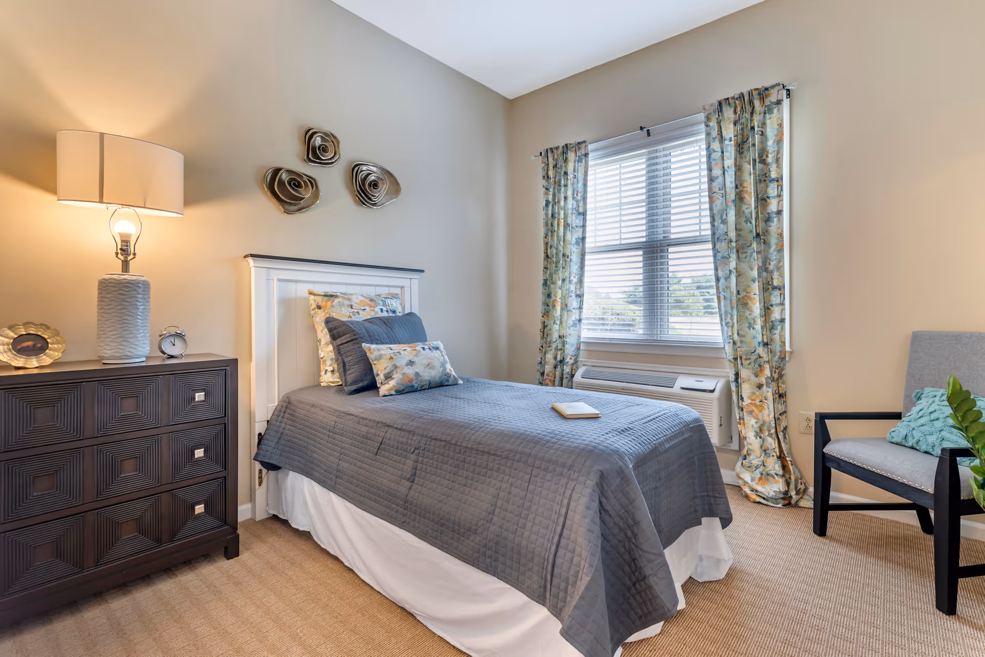 A neatly arranged bedroom with a single bed covered in a gray quilt and floral pillows. There is a dark wooden dresser with a lamp, a small clock, and a decorative item on it. The room has beige walls, a window with floral curtains, and a gray armchair with a green pillow beside the bed.