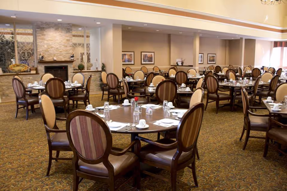 A spacious dining room with multiple round tables set with white cups, glasses, napkins, and silverware. The room features upholstered chairs with striped backs and a patterned carpet. The walls are decorated with framed pictures, and there is a stone fireplace with decorative items on the mantel.