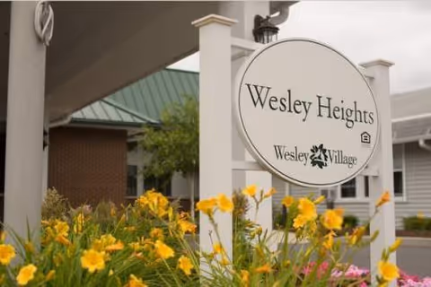 Outdoor view of the entrance sign for Wesley Heights at Wesley Village, surrounded by yellow flowers and greenery with buildings in the background.