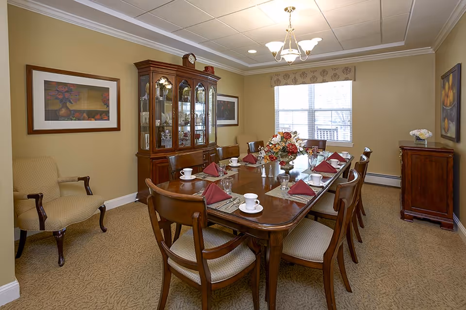 A formal dining room with a long wooden dining table set for eight people. The table is arranged with white cups, glasses, and burgundy napkins folded on placemats. A floral centerpiece decorates the middle of the table. The room has beige walls, carpeted floor, a window with a valance, two framed paintings on the walls, a wooden china cabinet displaying dishes, and a wooden sideboard with a small flower arrangement.