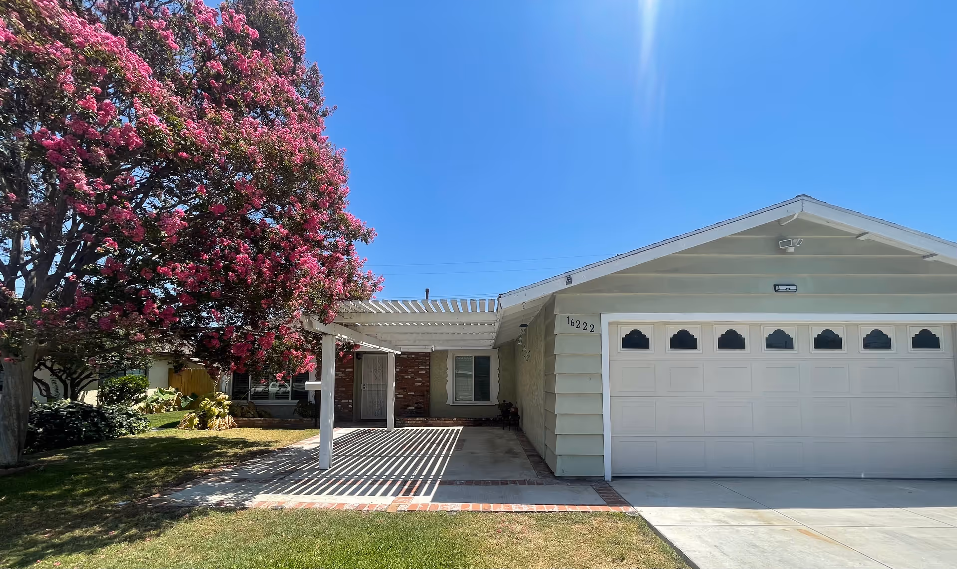 Front exterior view of a single-story house with a large blooming tree with pink flowers on the left side, a white pergola over the driveway, and a two-car garage with white doors under a clear blue sky.