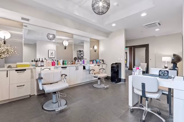 A modern salon area inside a senior living facility with two salon chairs in front of a large mirror and a counter filled with various hair care products. There is also a manicure station with a chair and nail care items. The room has bright lighting and a clean, spacious layout.
