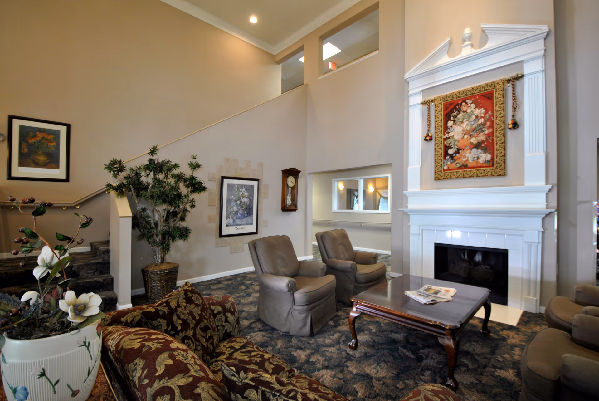 A cozy living room area in a retirement community featuring a patterned sofa, two armchairs, a wooden coffee table with newspapers, a white fireplace with a floral tapestry above it, framed artwork on the walls, a potted plant, and a staircase leading to an upper level.