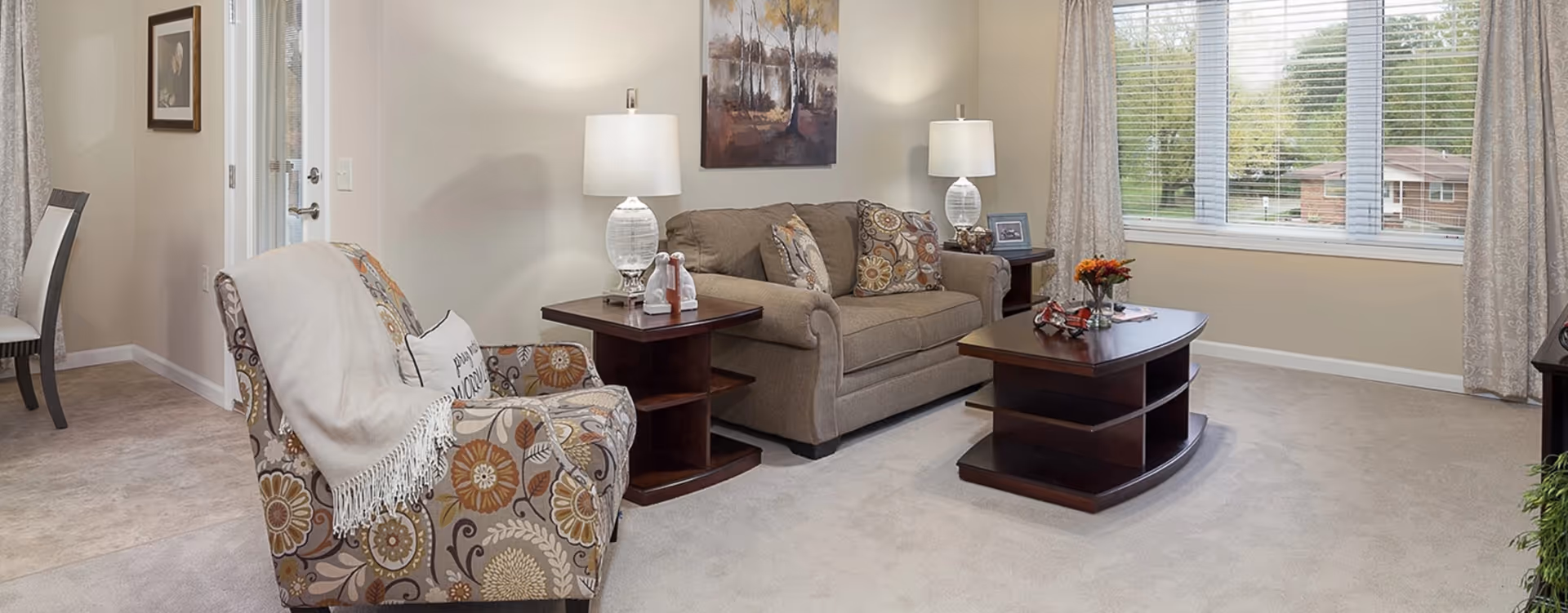 Bright living room with a sofa, patterned armchair, wooden coffee and side tables with lamps, and a large window overlooking trees.