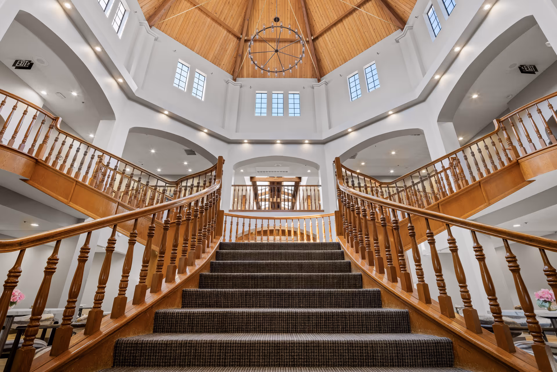 Carpeted staircase with wooden railings leading up to a spacious, well-lit interior area with a high wooden ceiling and large windows. The area features white walls, recessed lighting, and a large circular chandelier hanging from the ceiling.