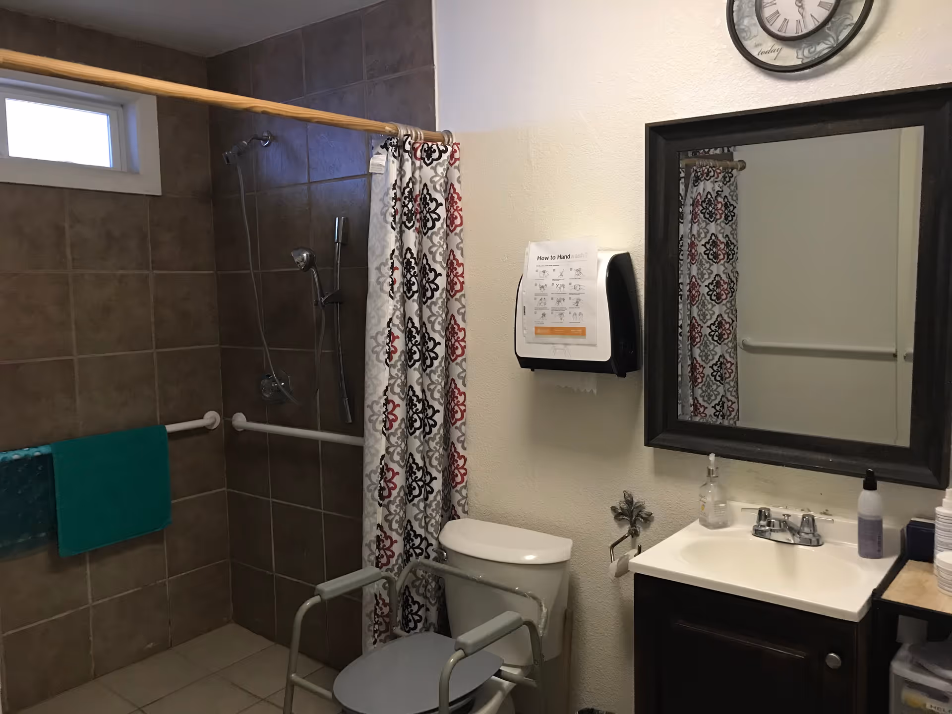 Bathroom with a tiled shower area featuring a handheld showerhead and grab bars. A shower curtain with a black, red, and gray pattern hangs on a wooden rod. There is a toilet with a raised seat and armrests, a sink with a mirror above it, a clock on the wall, and a paper towel dispenser with handwashing instructions.