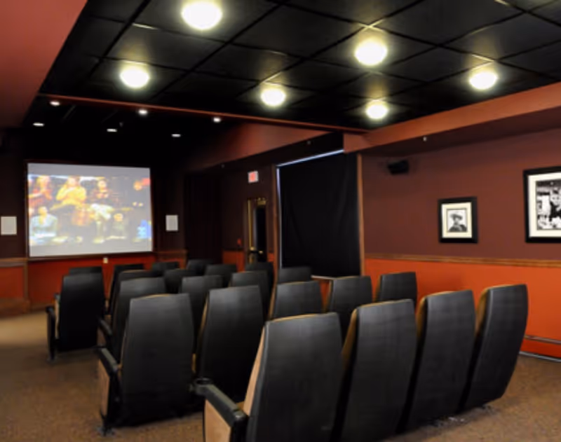 A small theater room with several rows of black cushioned seats facing a large screen showing a movie. The walls are painted dark red and decorated with framed pictures. The ceiling has recessed lighting.