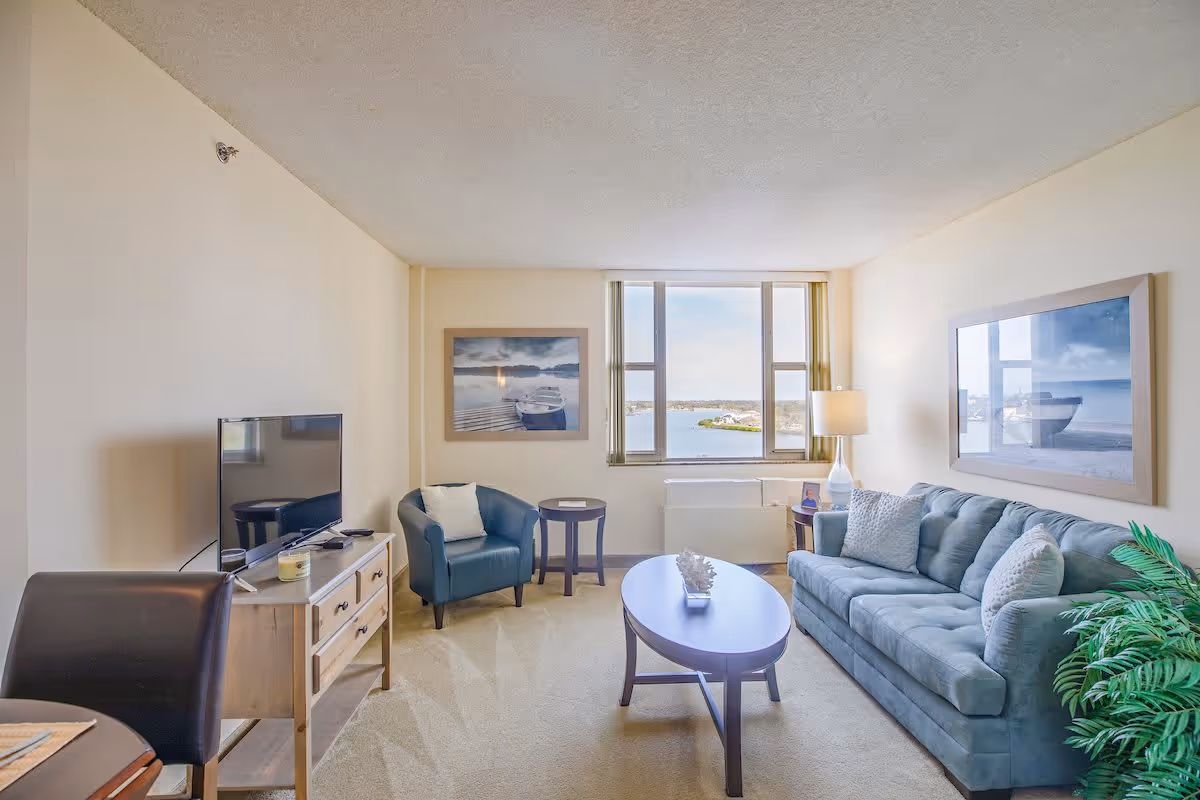 A bright living room with a large window showing a water view. The room features a blue sofa with cushions, a blue armchair, a wooden coffee table with a decorative item, a TV on a wooden stand, a floor lamp, and framed pictures of boats on the walls. There is also a green plant in the corner.